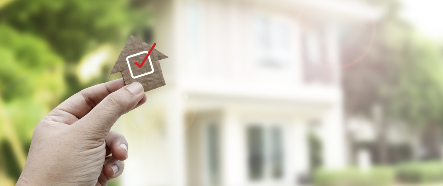 Hand holding a wooden house cutout with a red checkmark in front of a home